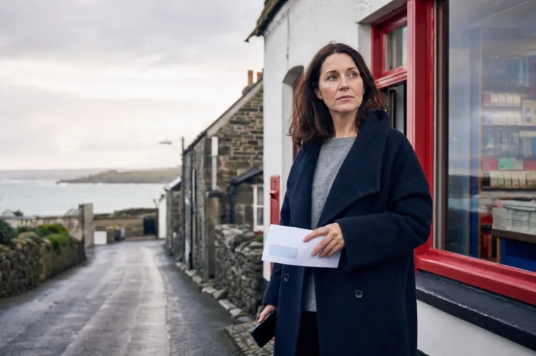 A thoughtful woman in rural Cornwall holding a cheque outside a small village post office on an overcast day.